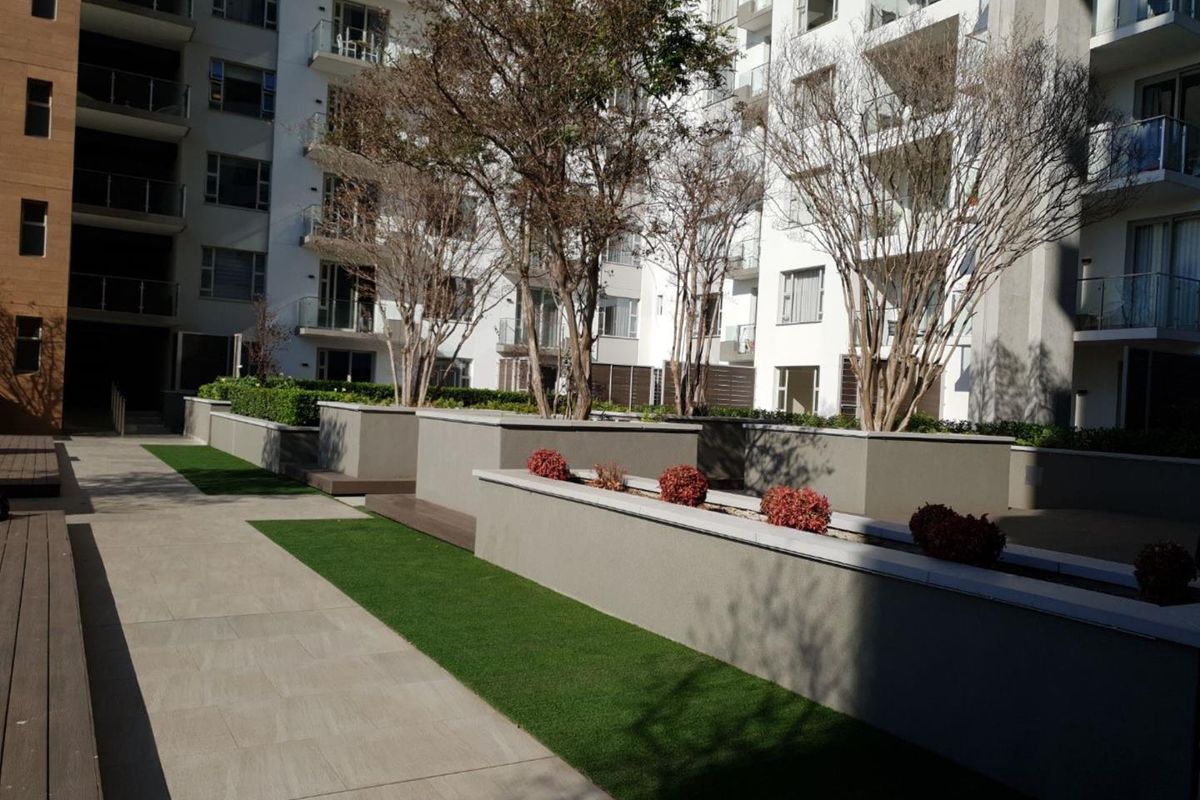 A well-maintained communal courtyard area featuring raised concrete planters, artificial turf, and tiled walkways set against a modern multi-story apartment building.