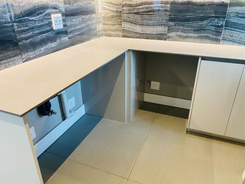 A clean, modern kitchen area featuring stone countertops, decorative tiled backsplash, and dedicated plumbing and electrical connections for appliances.