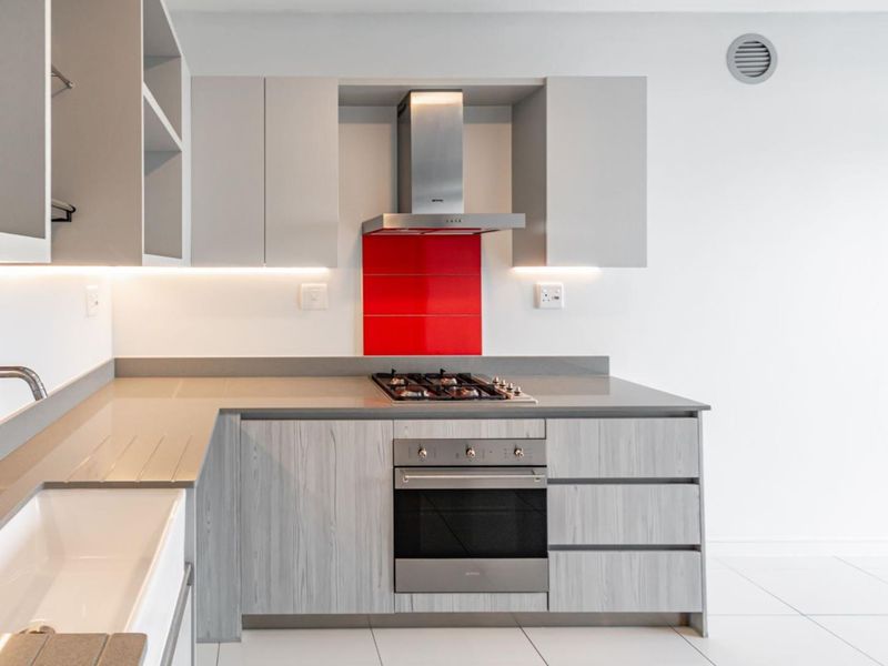 Modern kitchen featuring light wood-grain cabinetry, stone countertops, a built-in oven, gas hob, and a striking red glass splashback.