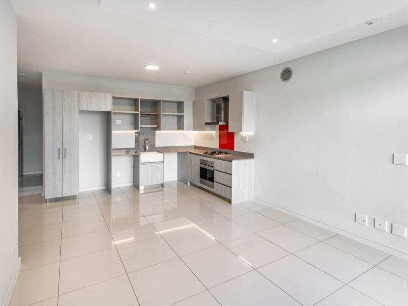 Modern open-plan kitchen featuring light wood-grain cabinetry, a red glass splashback, integrated gas hob, electric oven, and sleek ceramic floor tiles.
