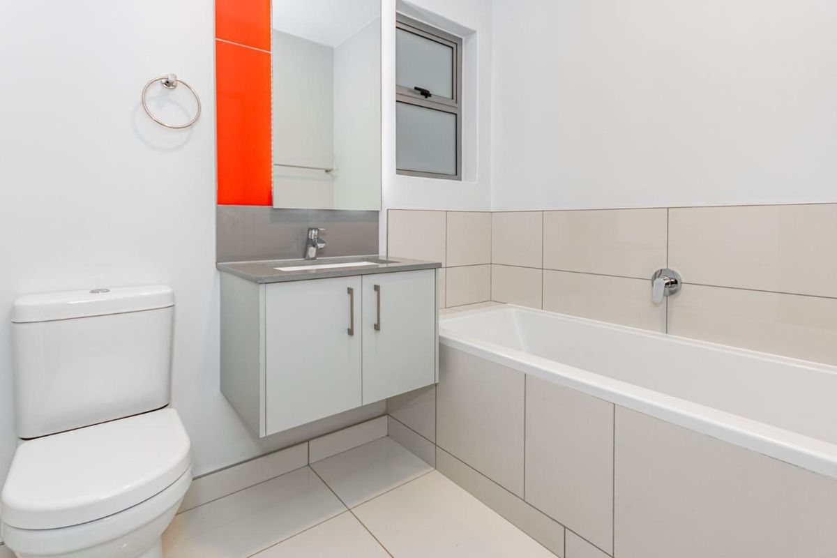 A clean, modern bathroom featuring a wall-mounted vanity, integrated basin, bathtub, and neutral floor-to-ceiling tiling with a vibrant orange accent panel.