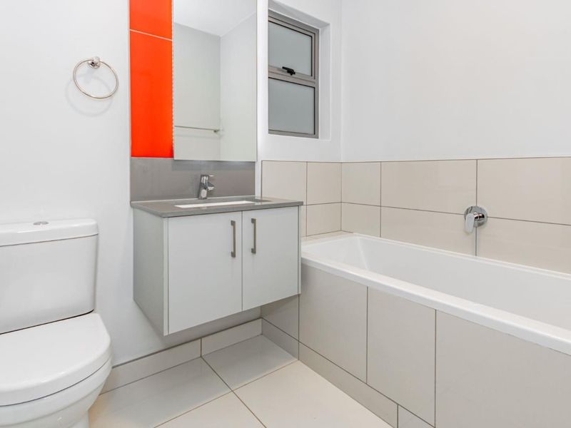 A clean, modern bathroom featuring a wall-mounted vanity, integrated basin, bathtub, and neutral floor-to-ceiling tiling with a vibrant orange accent panel.