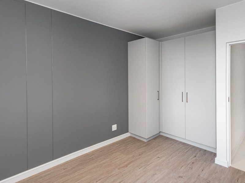 A clean, modern bedroom featuring neutral grey accent walls, light wood-look laminate flooring, and ample built-in cupboard storage.