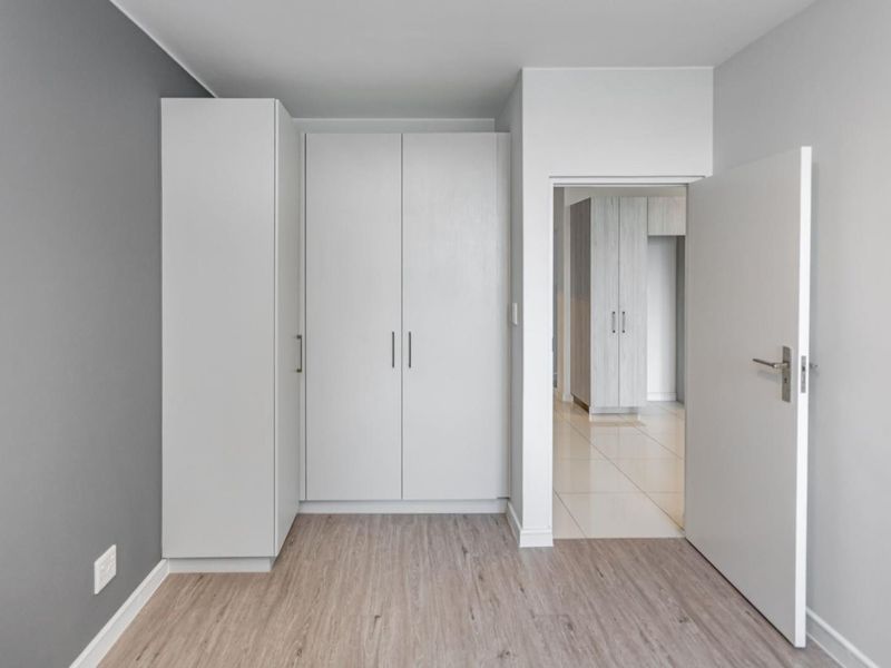 A clean, modern bedroom featuring built-in cupboards, light laminate flooring, and a neutral grey accent wall, leading into a tiled hallway.