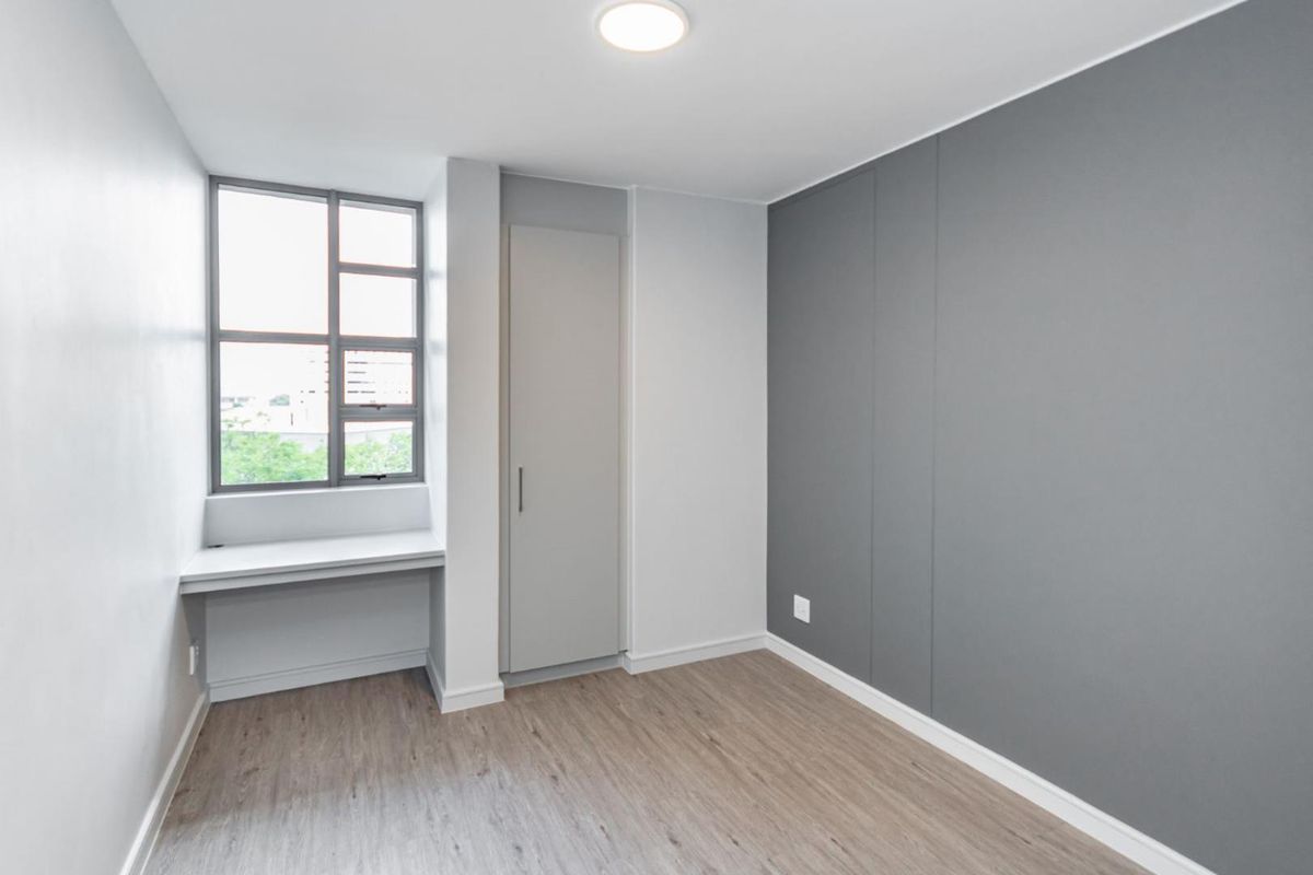 A clean, modern bedroom featuring neutral grey tones, laminate flooring, a built-in cupboard, and a convenient integrated desk space by the window.