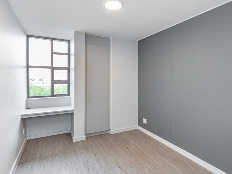 A clean, modern bedroom featuring neutral grey tones, laminate flooring, a built-in cupboard, and a convenient integrated desk space by the window.