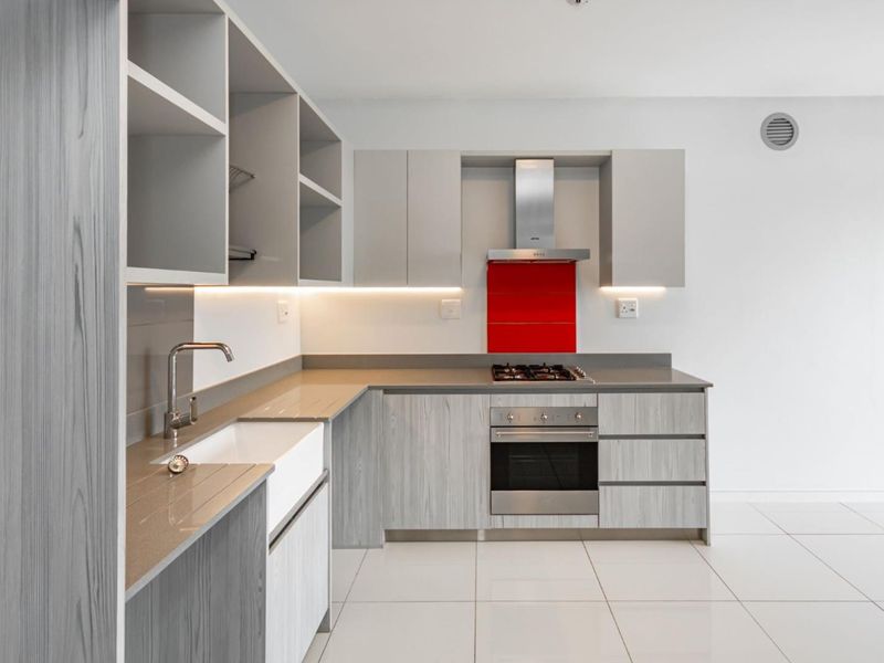 Modern kitchen featuring light wood-grain cabinetry, stone countertops, a farmhouse sink, and integrated stainless steel appliances with a vibrant red splashback.
