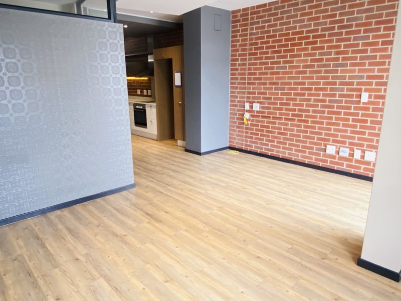 An open-plan living area featuring modern laminate flooring and an exposed brick feature wall, with a glimpse into the adjacent kitchen.