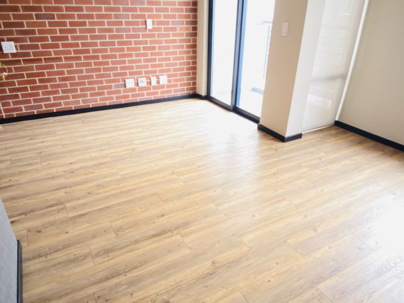 A bright, empty living area featuring a modern exposed brick feature wall, wood-look laminate flooring, and direct access to a balcony through sliding glass doors.
