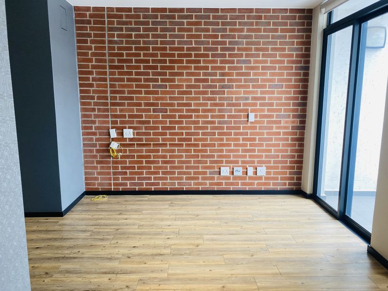 An empty living area featuring a prominent exposed brick wall, modern laminate flooring, and large sliding glass doors providing natural light.