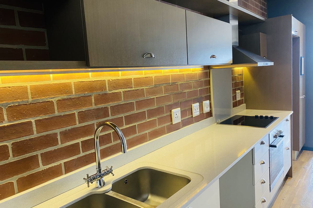 Modern kitchen featuring sleek cabinetry, stone countertops, integrated appliances, and a stylish brick splashback with ambient under-cabinet lighting.