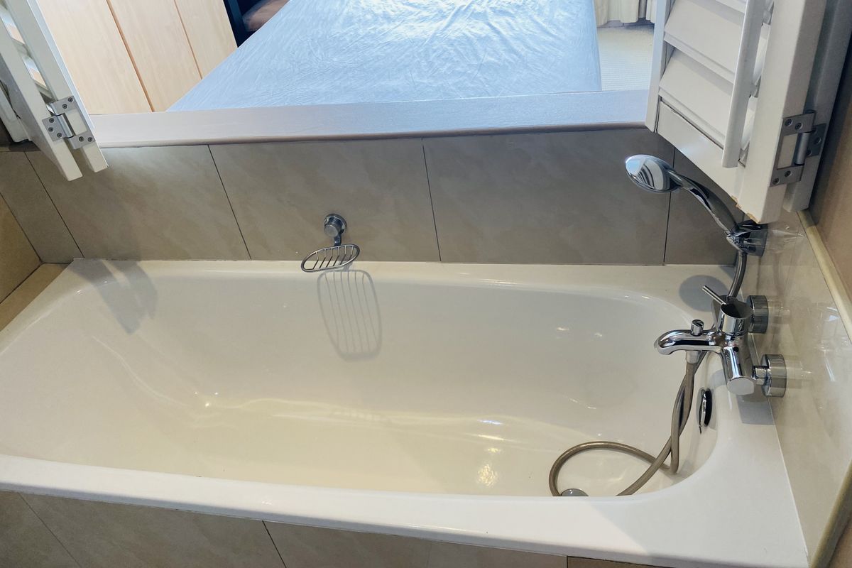 A bathroom featuring a built-in bathtub with chrome fixtures and a shuttered window opening into the adjacent bedroom.