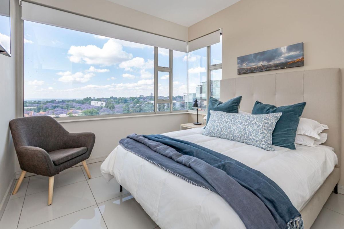 A bright bedroom featuring large corner windows with expansive city views, neutral walls, and modern tiled flooring.