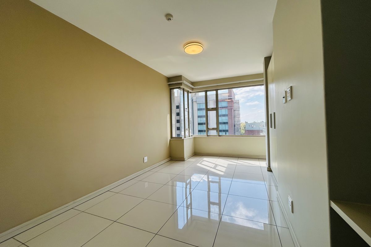 A bright, empty bedroom featuring polished tiled flooring, built-in cupboards, and large windows offering natural light and urban views.