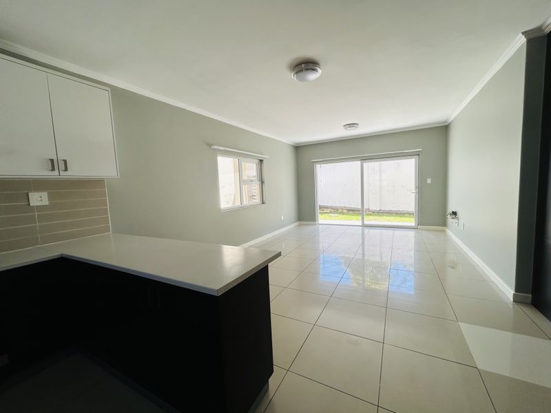 Bright open-plan living area featuring modern tiled flooring, neutral walls, and a functional kitchen with stone countertops, leading to a private outdoor space.