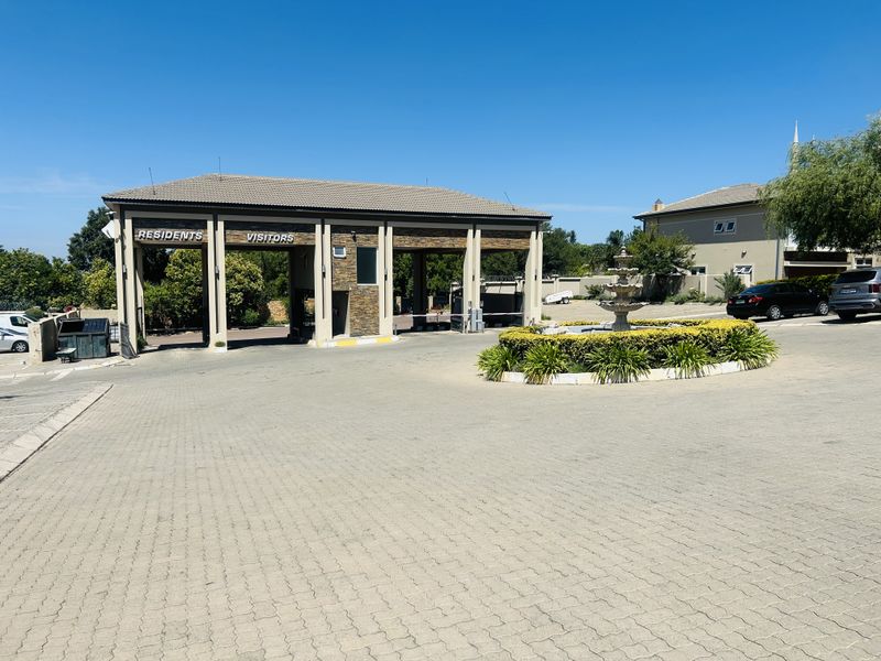 The entrance to the apartment complex features a secure gatehouse with separate lanes for residents and visitors, complemented by a paved driveway and fountain.