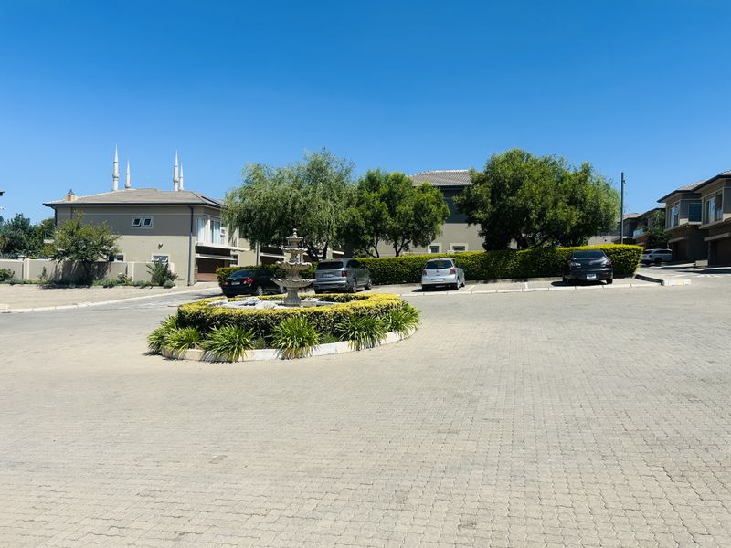 A well-maintained residential complex entrance featuring a paved driveway, central landscaped garden bed, and a decorative stone fountain.