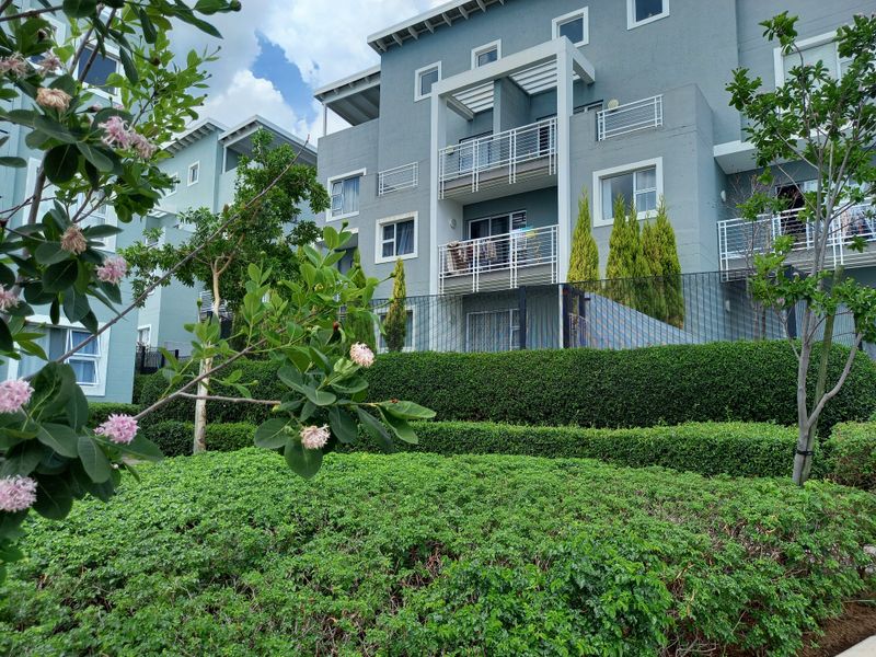 A view of the apartment building exterior featuring multiple balconies with metal railings, set behind well-maintained landscaped hedges and perimeter fencing.