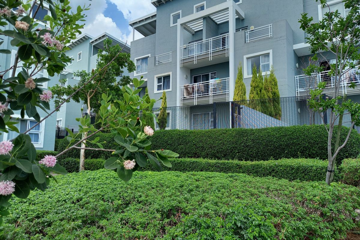 A view of the apartment building exterior featuring multiple balconies with metal railings, set behind well-maintained landscaped hedges and perimeter fencing.