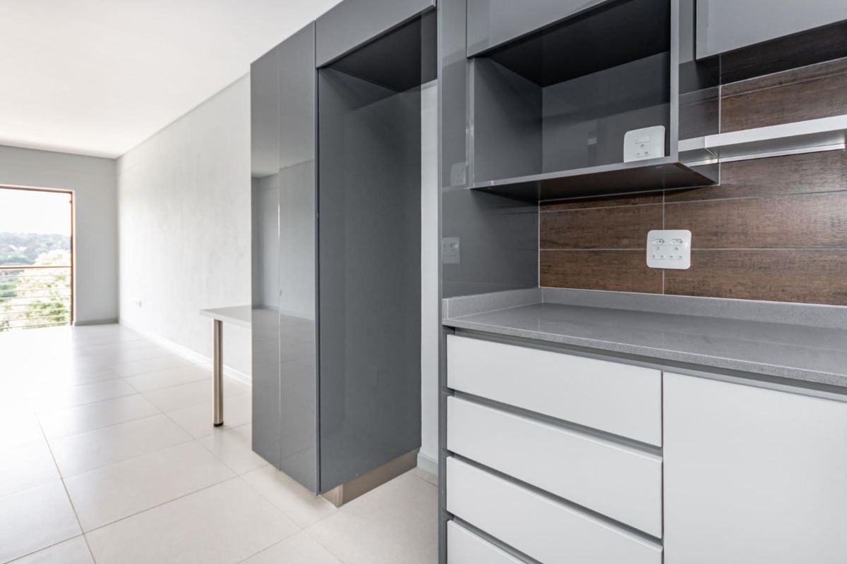 Modern kitchen area featuring sleek grey and white cabinetry, stone countertops, and tiled backsplash, leading to an open living space with balcony access.