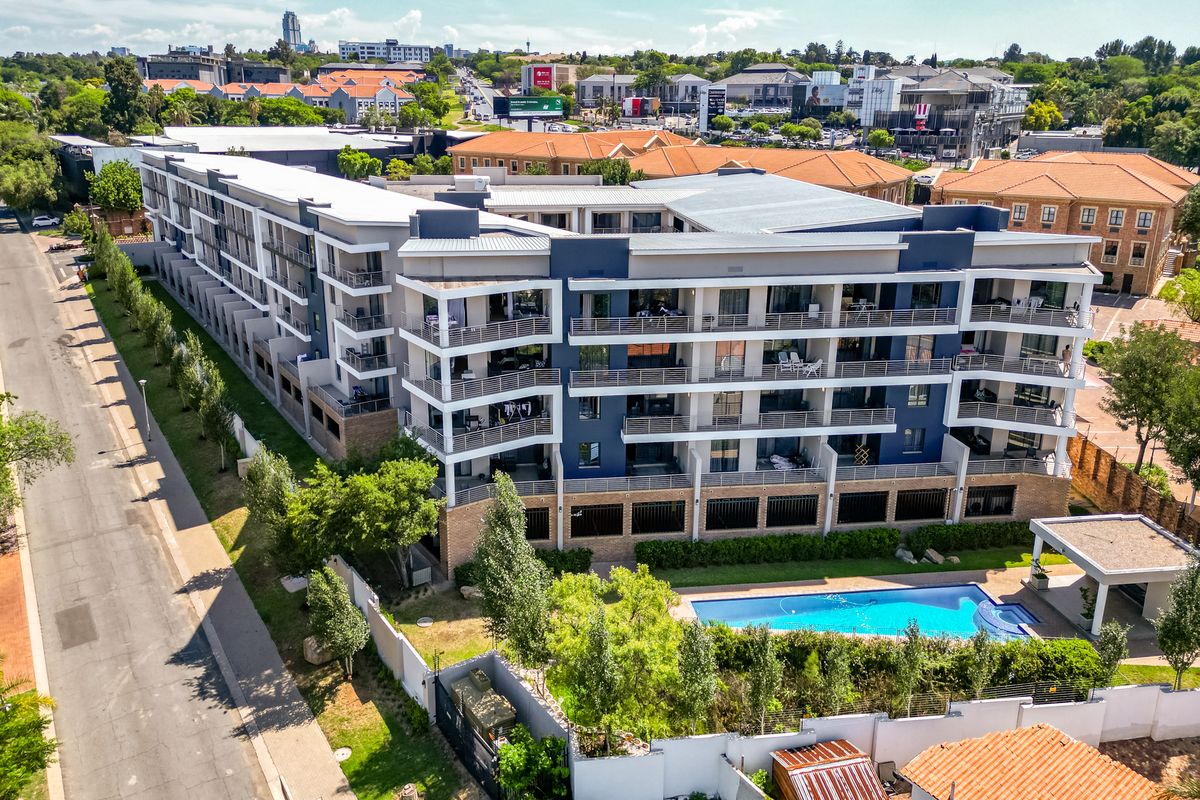 Aerial view of a modern multi-story apartment complex featuring private balconies, a swimming pool, and landscaped grounds in a suburban setting.