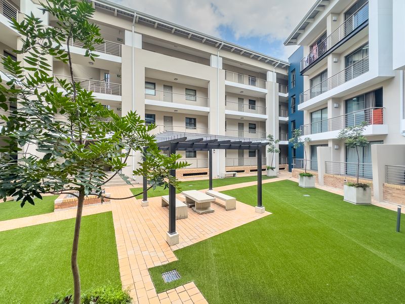 A well-maintained communal courtyard area featuring a pergola, paved seating space, and artificial lawn surrounded by modern apartment building facades.