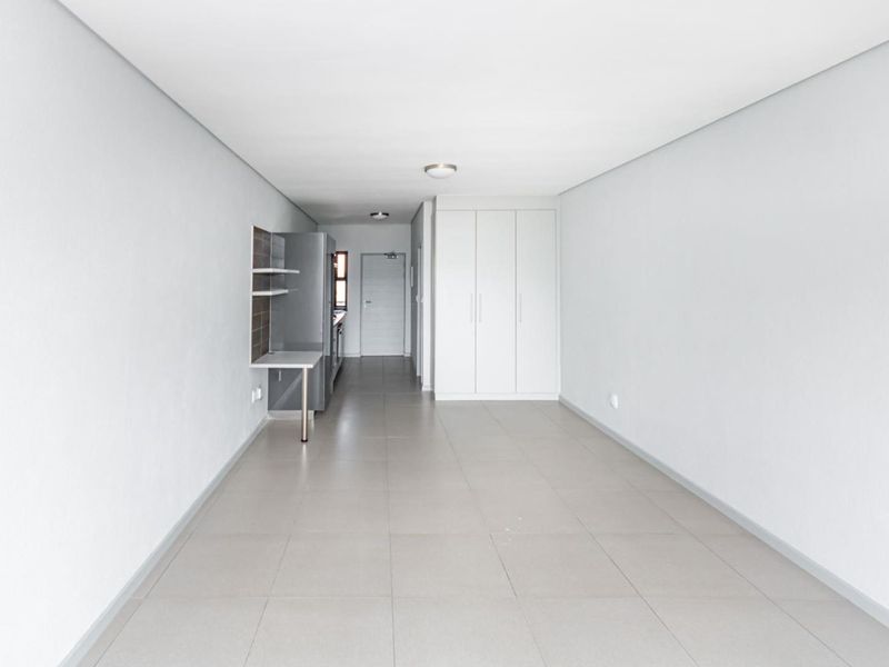 A bright, open-plan living area featuring neutral tiled flooring, built-in storage cupboards, and a compact kitchenette section.