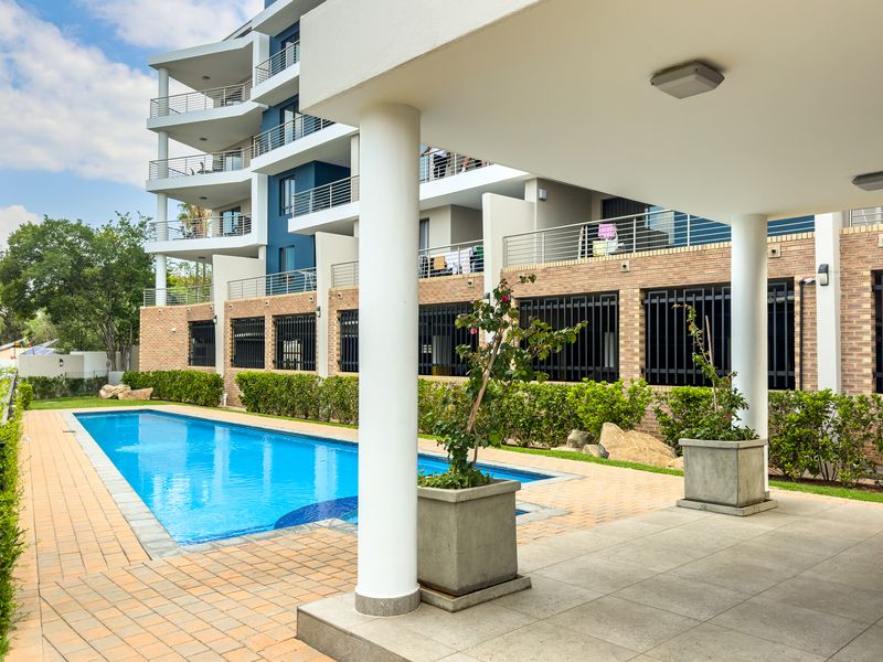A well-maintained apartment complex pool area featuring a paved deck, covered patio space, and landscaped garden borders.