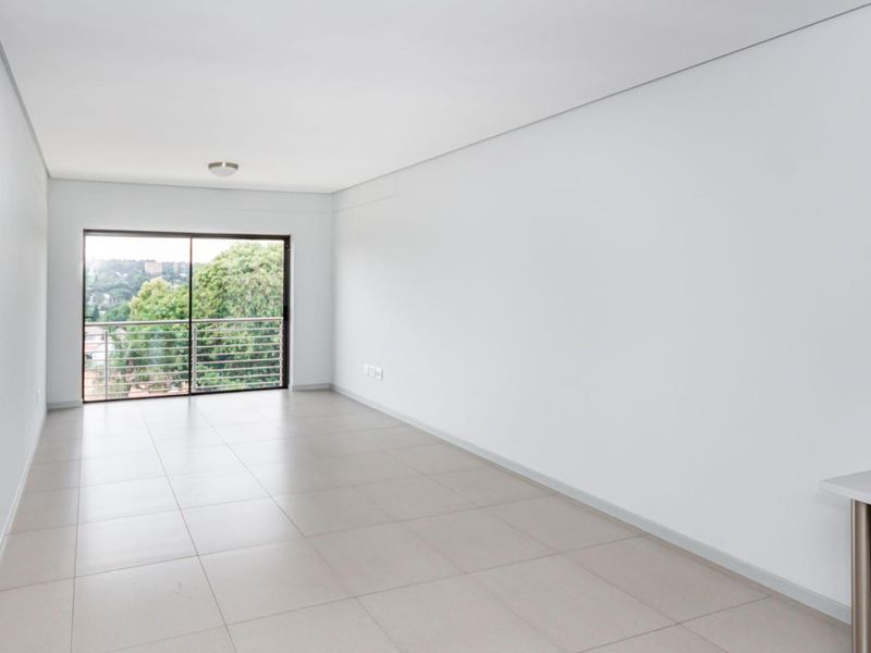 A bright, empty living area featuring neutral tiled flooring, clean white walls, and a sliding glass door leading to a balcony with scenic views.
