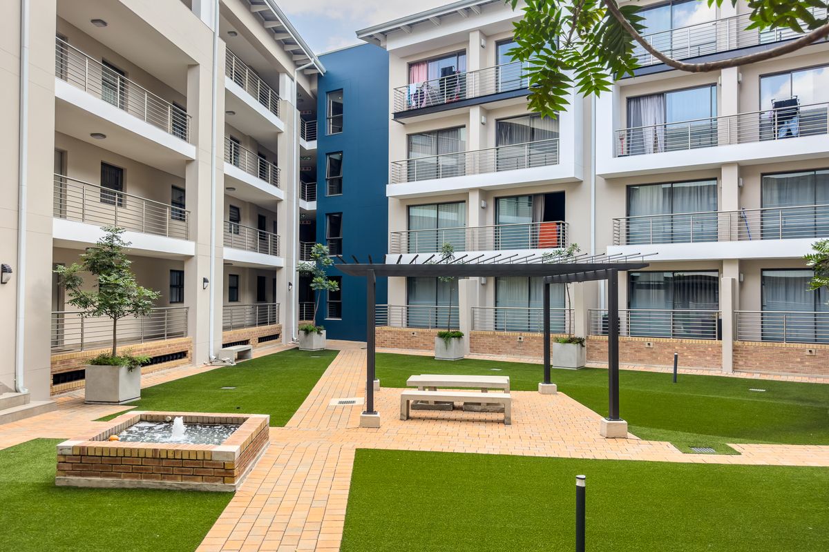 A well-maintained communal courtyard area featuring artificial lawn, brick pathways, a small water feature, and a pergola, surrounded by apartment building facades.
