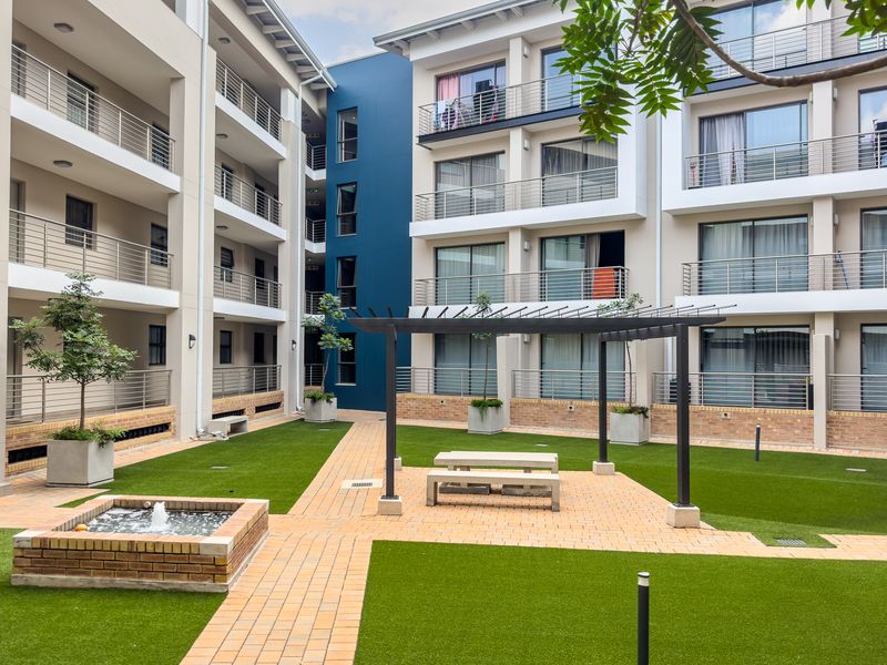 A well-maintained communal courtyard area featuring artificial lawn, brick pathways, a small water feature, and a pergola, surrounded by apartment building facades.