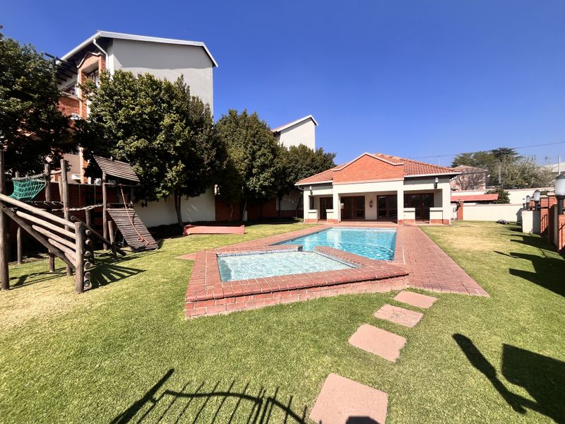Communal pool area featuring a swimming pool, integrated spa, brick paving, and access to a clubhouse within a landscaped garden setting.