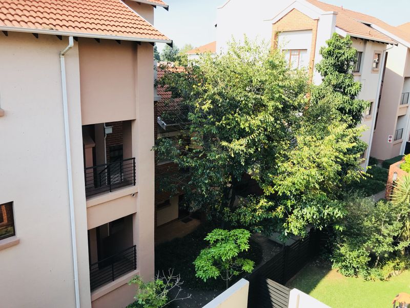 An elevated view of the apartment complex exterior, featuring multi-level balconies, terracotta roofing, and a well-maintained communal garden area.