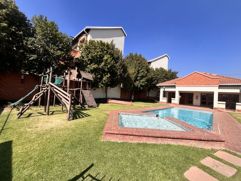 Outdoor communal area featuring a swimming pool with brick coping, paved surround, and a children's playground set within a landscaped garden.
