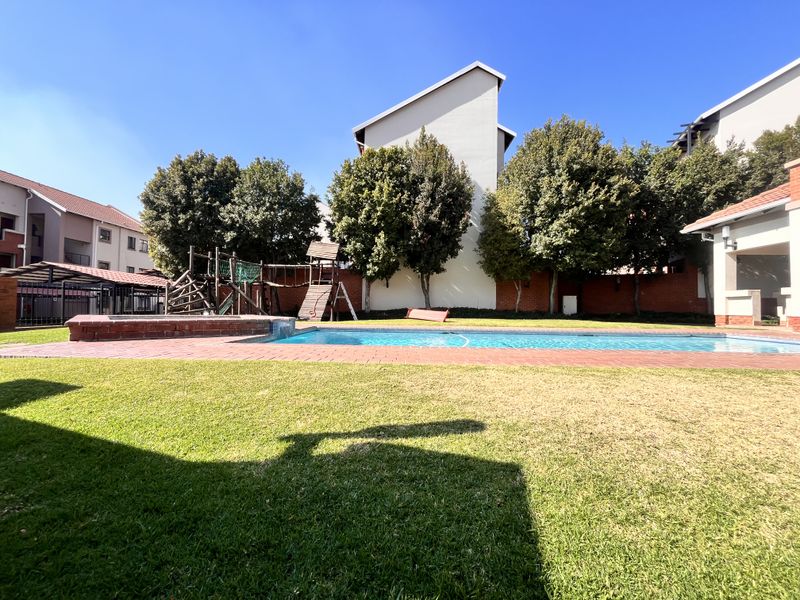Communal outdoor area featuring a swimming pool, brick paving, and playground equipment set within a landscaped garden environment.