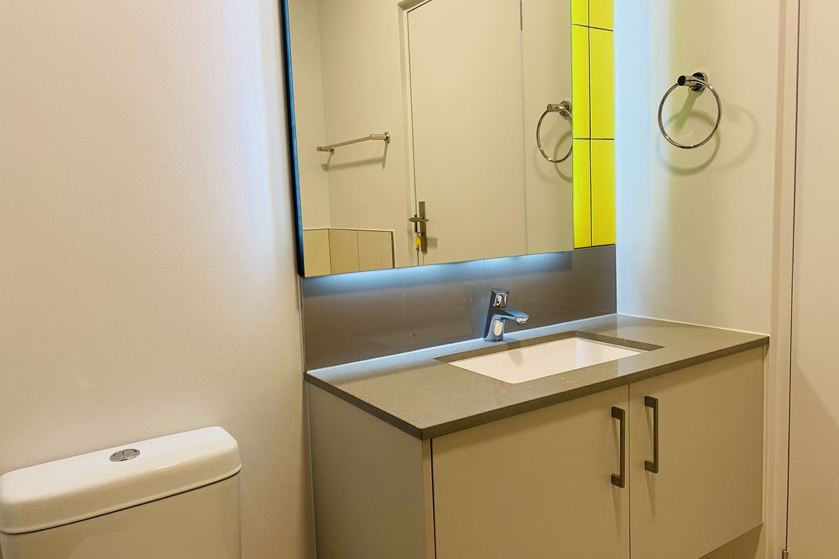 A modern bathroom featuring a floating vanity with stone countertop, undermount sink, backlit mirror, and a bright yellow tiled accent wall.
