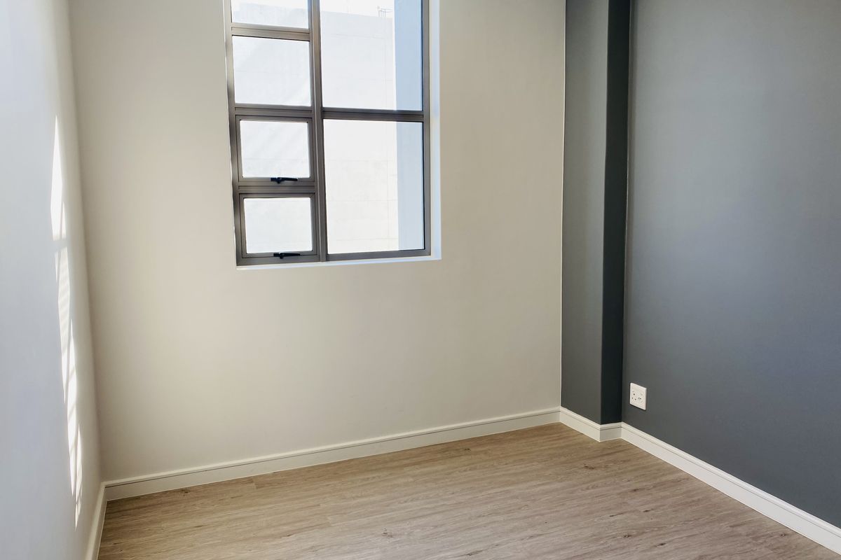 A bright, empty bedroom featuring modern laminate flooring, a neutral color palette with a dark grey accent wall, and an aluminium-framed window.