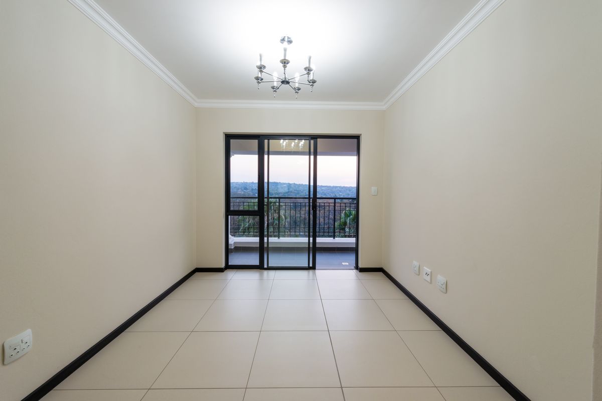 A bright, empty living room featuring neutral tiled flooring, crown molding, and sliding glass doors that open onto a balcony with scenic views.
