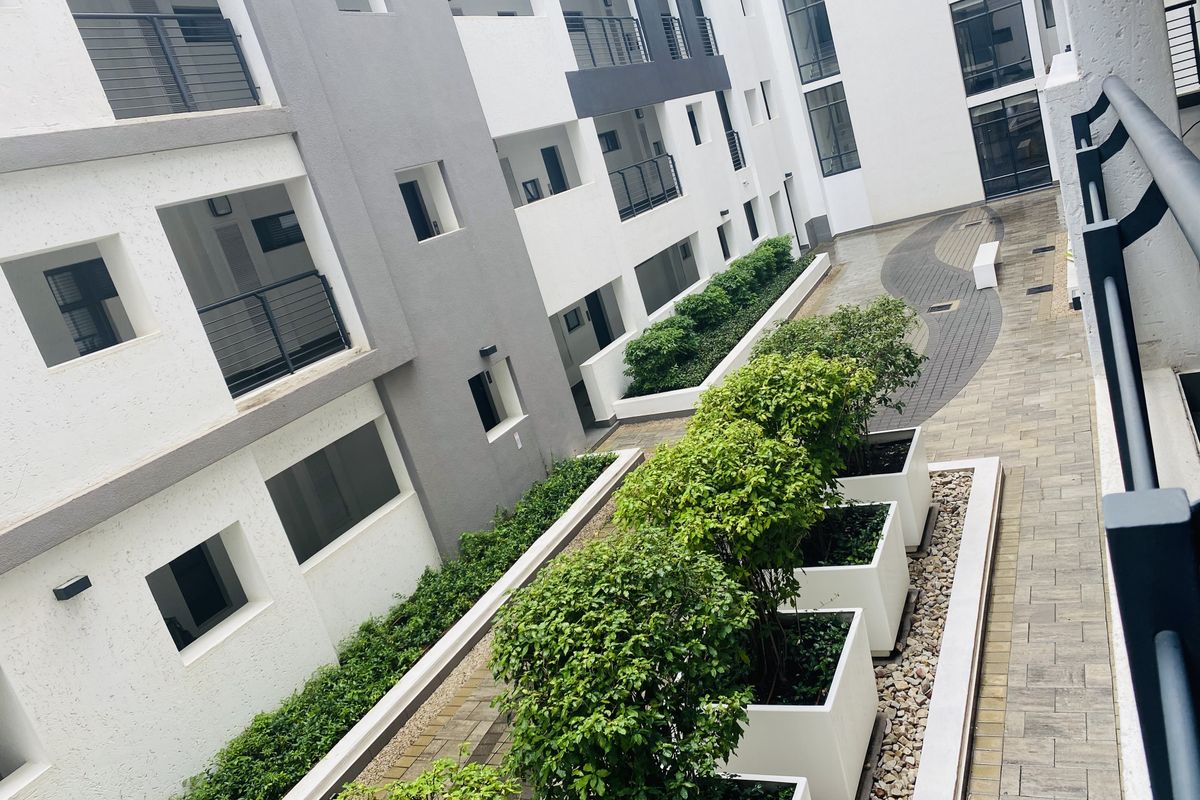 A modern apartment complex courtyard featuring paved walkways, raised garden planters, and multiple levels of balconies with metal railings.