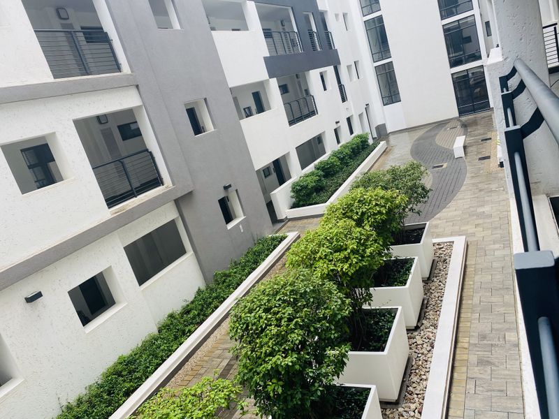 A modern apartment complex courtyard featuring paved walkways, raised garden planters, and multiple levels of balconies with metal railings.