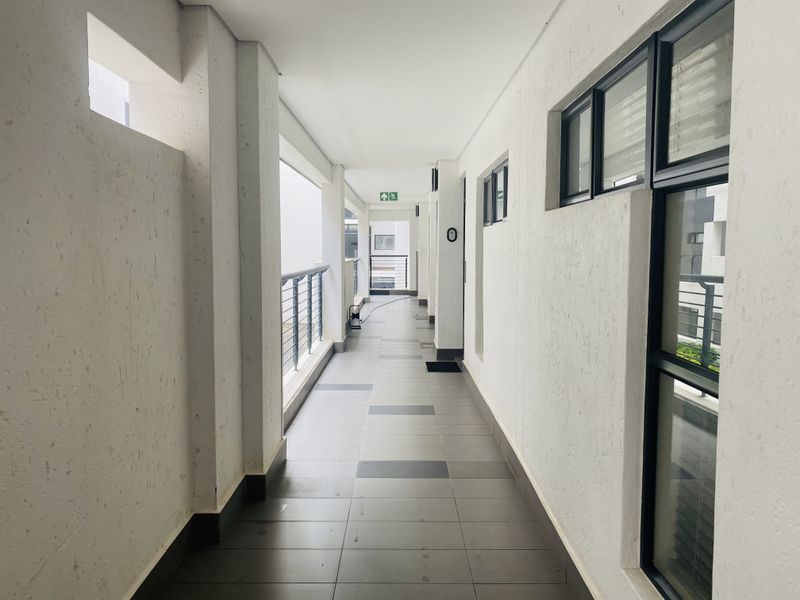 A clean, well-lit apartment hallway featuring tiled flooring, aluminium window frames, and a metal balustrade overlooking the building exterior.