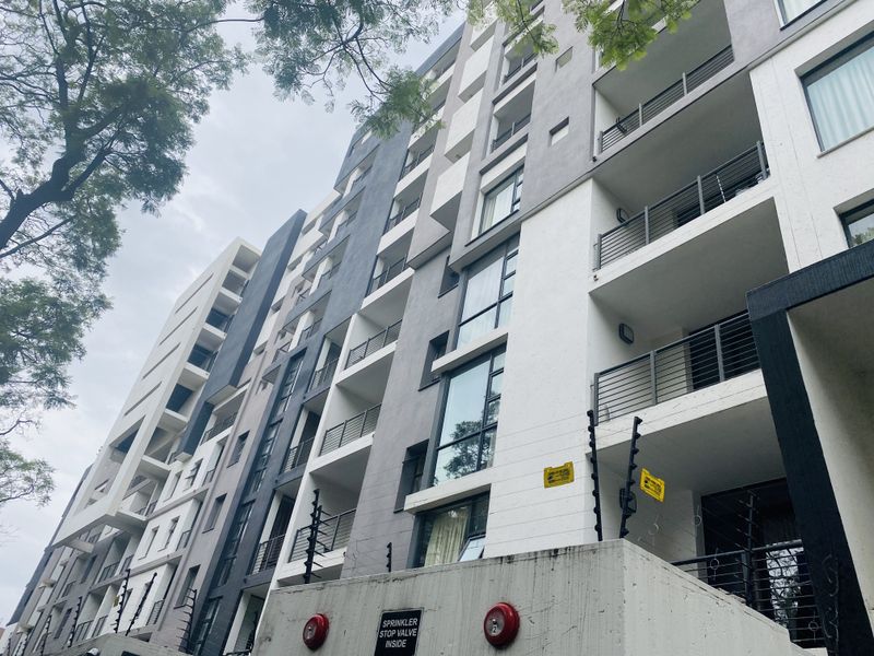 A low-angle view of a modern multi-story apartment building featuring balconies with metal railings, concrete facade elements, and integrated security fencing.