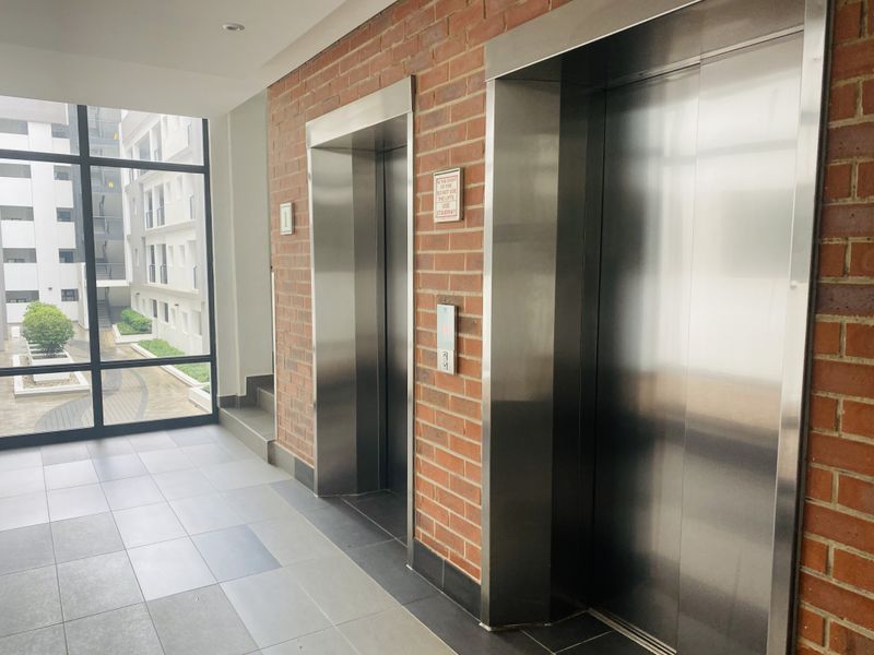 A modern apartment building hallway featuring stainless steel elevators, exposed brick wall detailing, and large windows overlooking the complex courtyard.