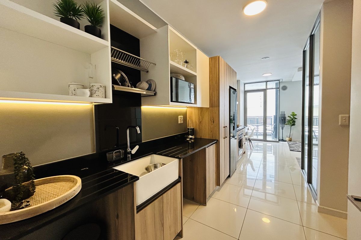Modern galley-style kitchen featuring sleek wood-tone cabinetry, black stone countertops, an apron-front sink, and integrated appliances, leading to a bright living area.