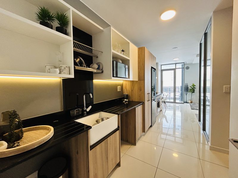 Modern galley-style kitchen featuring sleek wood-tone cabinetry, black stone countertops, an apron-front sink, and integrated appliances, leading to a bright living area.