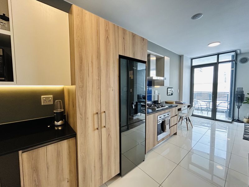 Modern open-plan kitchen featuring wood-grain cabinetry, stone countertops, integrated appliances, and seamless access to a private balcony through glass sliding doors.