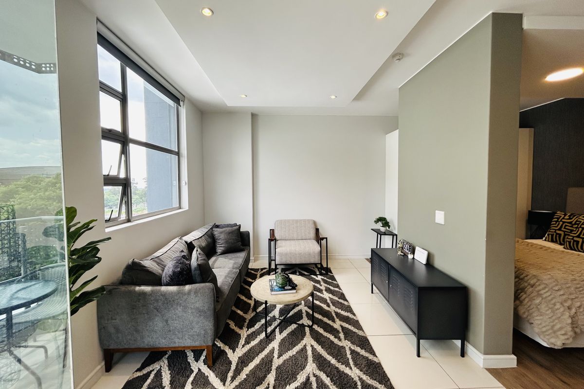 A bright and modern living area featuring neutral tiled floors, recessed ceiling lighting, and a partition wall separating the space from the bedroom.