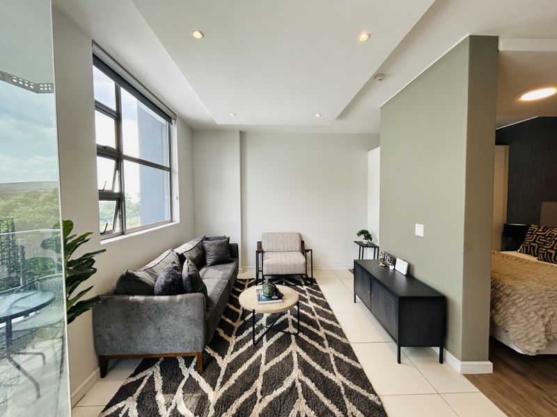 A bright and modern living area featuring neutral tiled floors, recessed ceiling lighting, and a partition wall separating the space from the bedroom.