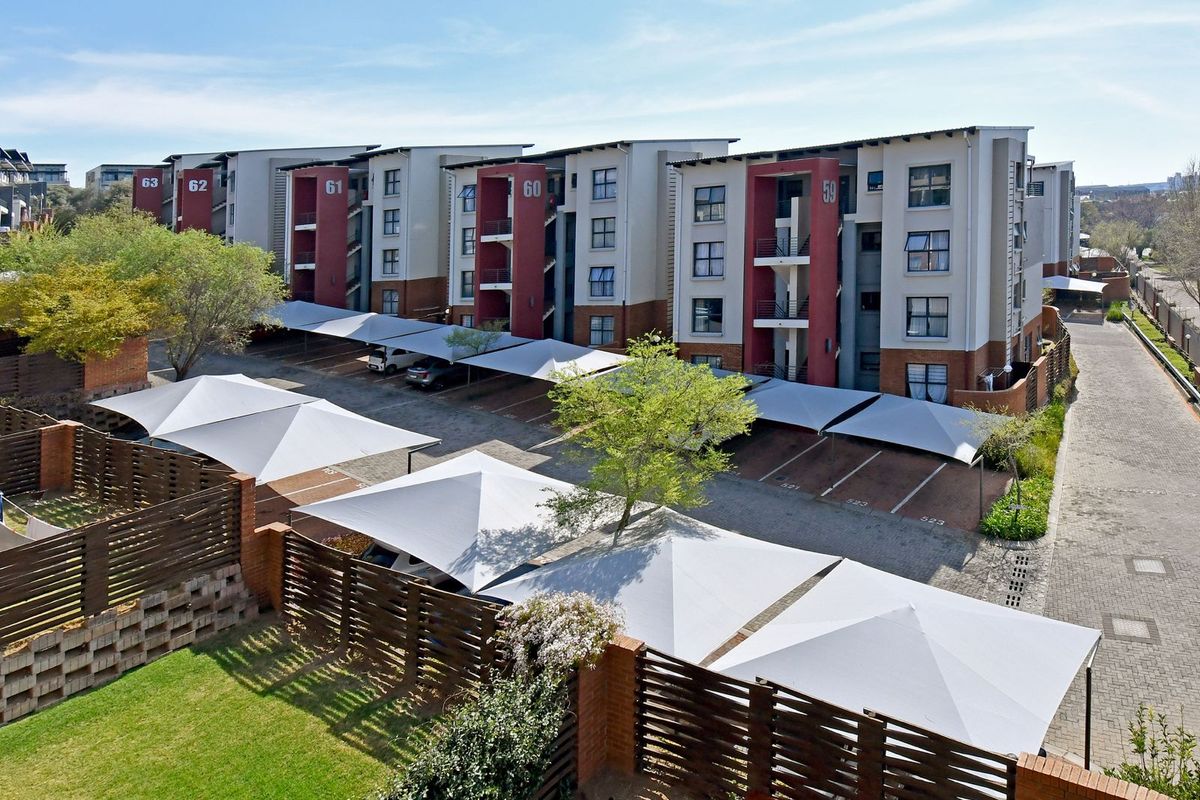 Aerial view of a modern apartment complex featuring multiple residential blocks, paved parking areas with shade cloth carports, and perimeter brick walling.