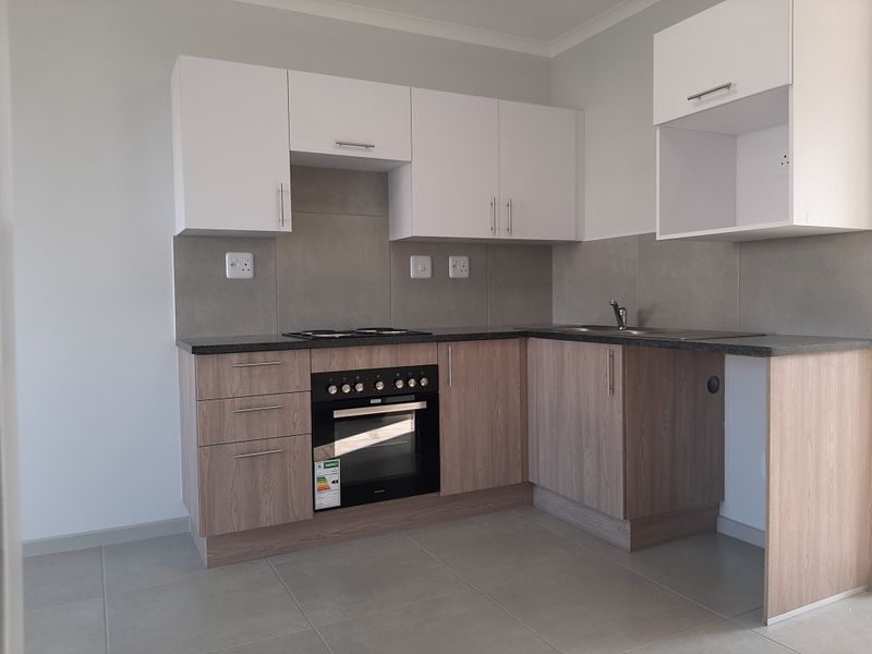 A clean, modern kitchen featuring light wood-finish cabinetry, white upper cabinets, dark countertops, and integrated electric oven and hob with tiled splashbacks.